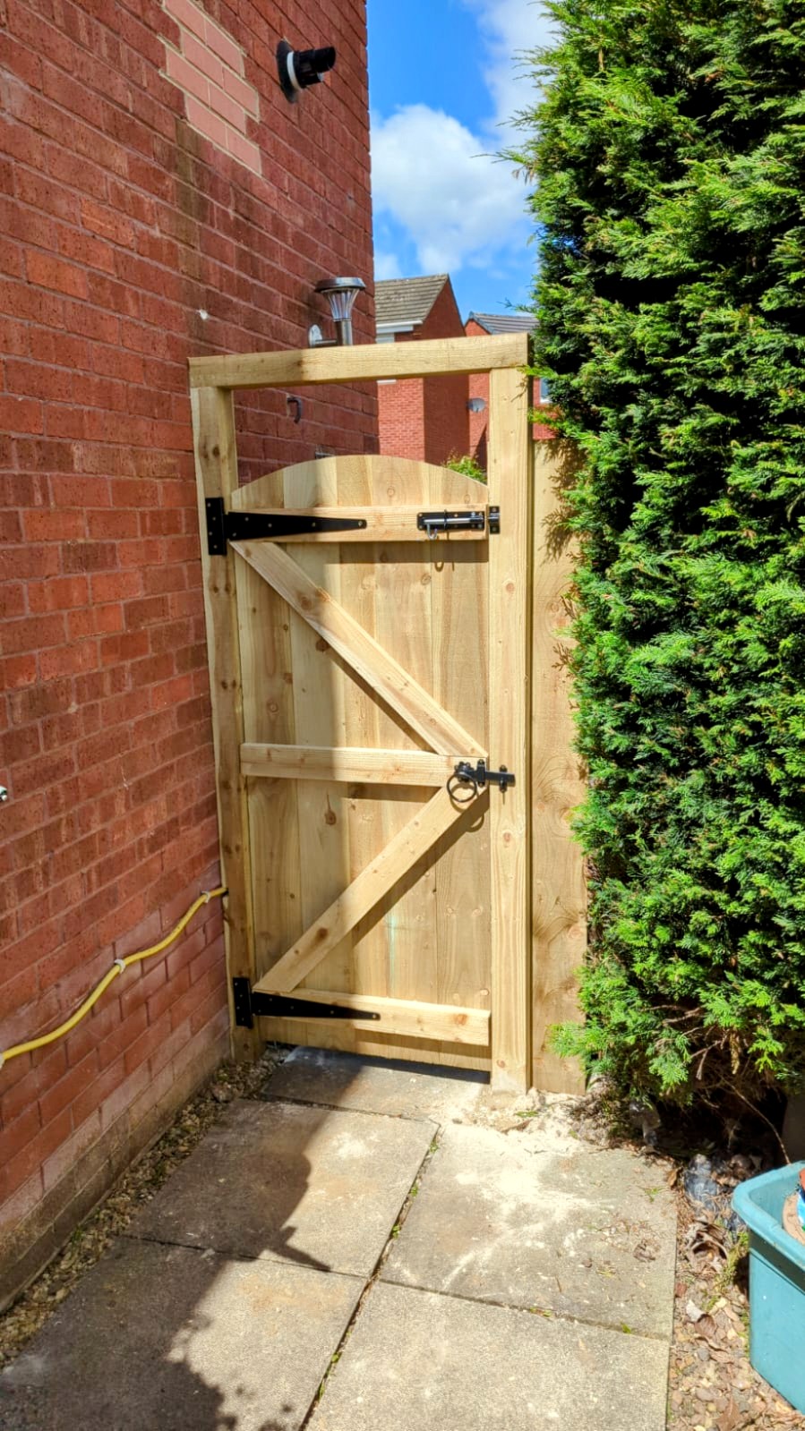 A ledged timber side gate with black hinges between a brick wall and a hedge